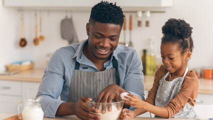A father and his daughter are having fun in the kitchen. They mix ingredients in a bowl. The girl...