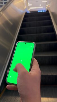Hand Holding Smartphone with Green Screen on Escalator in Mall Vertical Video. Close-up of a person holding a smartphone with green screen chroma key while riding an escalator indoors.
