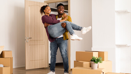 A young couple stands in a new apartment filled with moving boxes. The man lifts the woman playfully while they smile. They enjoy a joyful moment in their new home together. © Prostock-studio