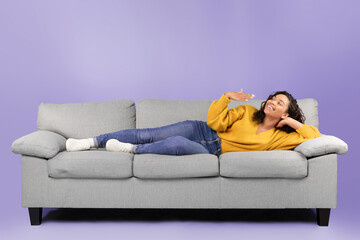 Obraz premium A woman with curly hair is lying on a gray couch. She wears a yellow sweater and blue jeans. She is smiling and looking at the camera while sitting comfortably.
