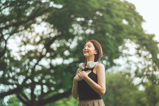 Young Asian woman practicing mindfulness and deep breathing in a lush green park under large trees. Wearing headphones, she enjoys a peaceful moment of mental wellness and nature connection