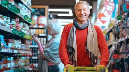A man stands with a shopping cart in a grocery store. He looks happy as he smiles at the camera. A woman is shopping in the background, checking items on the shelves. © Prostock-studio
