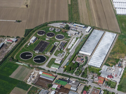 Aerial view of a wastewater treatment plant with circular sedimentation tanks and rectangular basins surrounded by greenhouses and agricultural fields in Paestum, Campania, Italy.