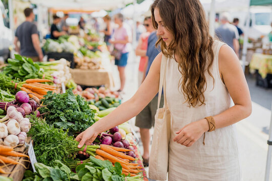 A young Caucasian woman in a light linen dress picks a fresh organic carrots at a sunny busy outdoor farmers market with vegetables like garlic and kale on display