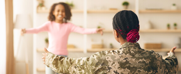 A soldier in military uniform stands with arms open wide, welcoming a smiling child in a pink shirt in a living room. The setting shows a warm environment with plants and shelves. © Prostock-studio