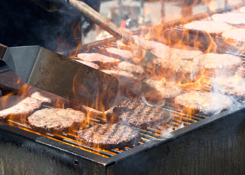 Close up on many hamburger patties, raw and cooked on an open BBQ grill outside, flames shooting up between the patties.
