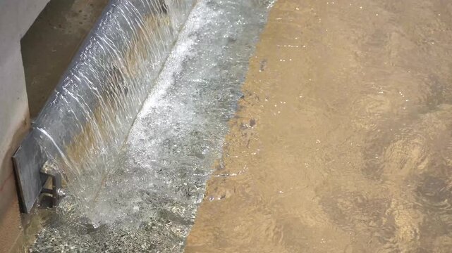 High-angle close-up of water cascading over a metal weir into a basin, creating bubbles and aeration. Ideal industrial B-roll for water treatment, fluid dynamics, and environmental engineering.