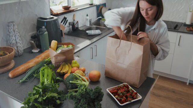 Young adult woman opens a brown paper bag in a kitchen filled with fresh fruits and vegetables. She sorts the groceries on the counter.
