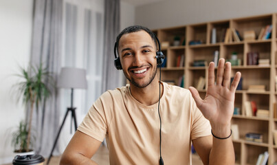 Cheerful arab man in headset having web conference, waving hand at camera, gesturing hello and smiling, enjoying online communication, webcam view © Prostock-studio