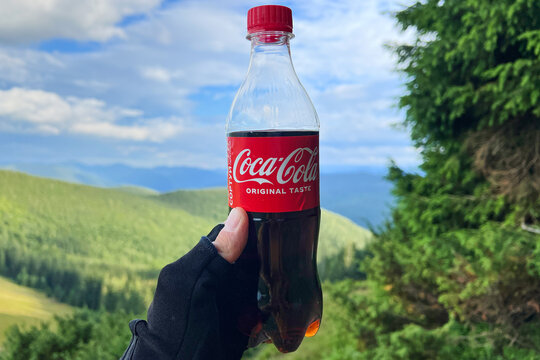 Hand holding Coca-Cola bottle on mountain trail overlook with blurred valley view. Outdoor beverage illustrative editorial stock photo: Bukovel, Ukraine - July 28, 2023