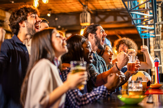 Friends cheering during soccer match in pub with beers on table.