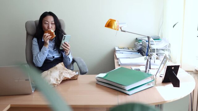 Asian woman worker in office having fast food lunch at desk. Woman eating hamburger while working in office in front of the computer.