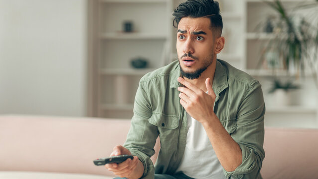 A man sits on a couch in a living room looking surprised while holding a remote control. He is focused on the screen ahead of him. The room has simple decor and soft lighting.