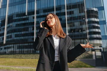 Fototapeta premium Beautiful Business Woman Talking On Smartphone And Gesturing Near Modern Office Building Wearing Black Suit. Corporate Lifestyle Concept Female Professional