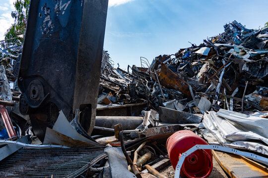 Hydraulic scrap shear machine at an industrial metal recycling yard surrounded by scrap metal and steel waste under blue sky.