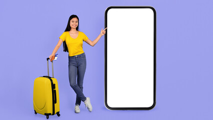 A young woman stands next to a large blank screen holding a suitcase. She smiles and points at the screen, suggesting travel plans or an upcoming event. © Prostock-studio