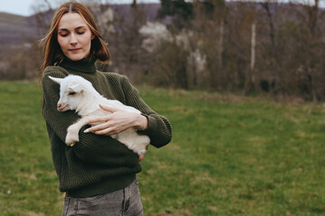 Fototapeta premium Calm woman outdoors holding a goat kid close, gentle expression, olive green sweater, soft natural light, rural field backdrop, relaxed posture, warm tones, candid lifestyle moment, fresh countryside