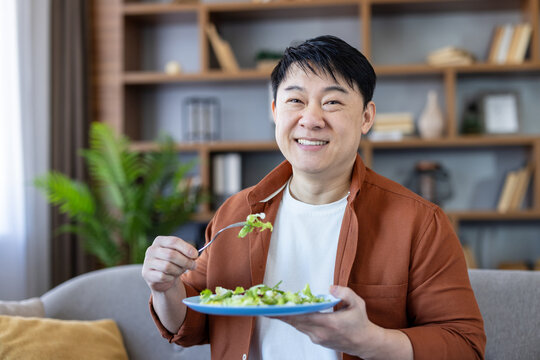 Happy asian man smiling at camera, holding a blue plate of fresh green salad at home, embracing mindful, plant-based eating and everyday wellness in a cozy living room