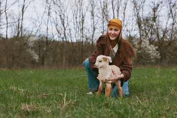 Fototapeta premium Cheerful woman outdoors with a goat kid, kneeling in a grassy field, warm brown jacket, light, candid lifestyle moment, gentle interaction, natural colors, tranquil countryside backdrop