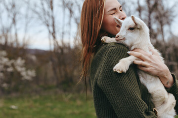 Fototapeta premium A woman in a green sweater cradles a young goat, her smile warm as they share a tender moment outdoors. Lifestyle scene emphasizing bond between human and animal in nature.