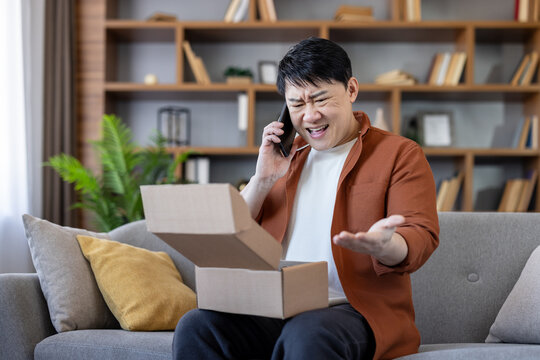 Asian man sitting on sofa at home, opening an empty or incorrect package box and making an angry, disappointed phone call to customer support about a delivery problem or an online order mistake