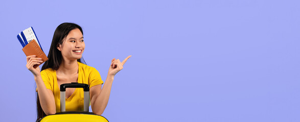 A woman stands with a yellow suitcase, holding travel tickets and a passport. She smiles and points to the side. The background is a solid color, creating a cheerful travel theme. © Prostock-studio
