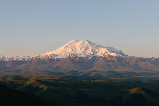 Snow-capped Mount Elbrus glowing at sunrise, viewed from Bermamyt Plateau with rolling hills and clear sky, North Caucasus, Karachay-Cherkessia, Russia