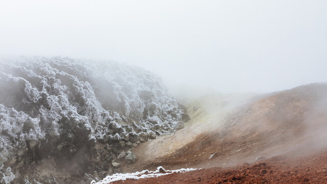 Steaming volcanic crater with snow-covered rocks and red ash slopes in Avachinsky Volcano, Kamchatka Peninsula, Russia, showcasing geothermal activity and rugged terrain