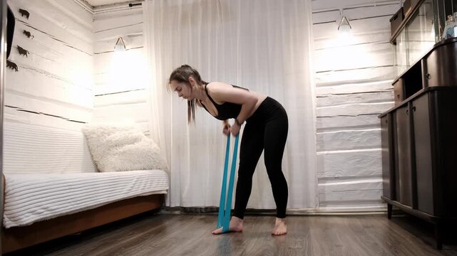 Caucasian woman doing exercise with an elastic band at home. Girl wearing black leggins and sports bra. Pilates workout. Strengthening the muscles of back, arms and shoulder girdle. 4k footage.