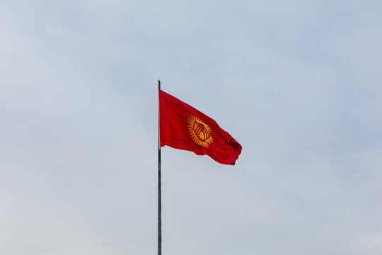 Red Kyrgyzstan national flag with sun and tunduk emblem waving on a tall flagpole against cloudy sky at Ala-Too Square, Bishkek, Chuy Region, Kyrgyzstan