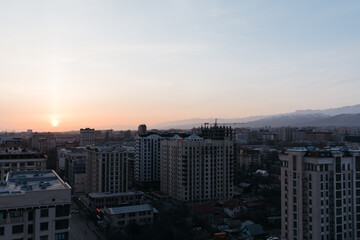 Obraz premium Wide cityscape view of Bishkek at sunset, featuring modern residential high rises, urban streets, and distant mountain range under a soft evening sky