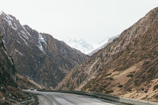 Winding highway through steep rocky mountains at Too Ashuu Pass, Chuy Region, Kyrgyzstan, with distant snow-covered peaks and a car on an overcast day