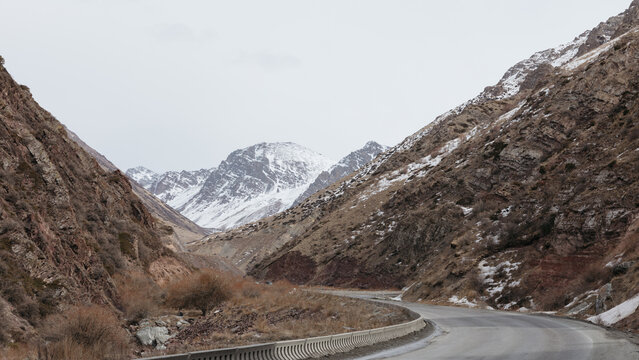 Scenic winding highway runs through a rugged mountain gorge with snowy peaks and rocky slopes at Too Ashuu Pass, Chuy Region, Kyrgyzstan under cloudy sky