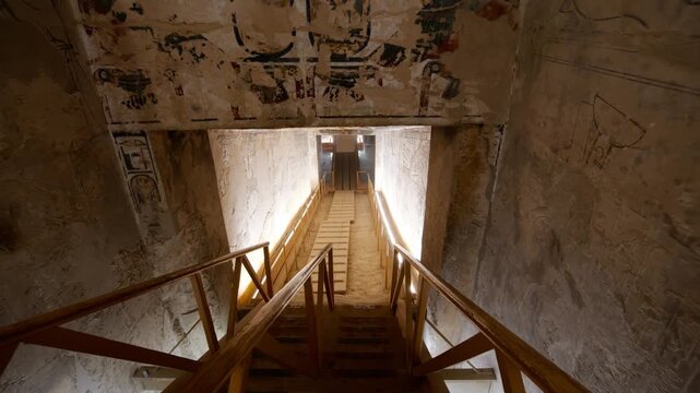 Narrow descending staircase inside an ancient Egyptian royal tomb, KV17 tomb of Seti I, Valley of the Kings, Luxor, Egypt, with faded hieroglyphic on ceiling, illuminated stone walls and wooden railin
