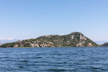 Naklejka premium Wide view of a rocky green island and calm rippled water on Lake Skadar in Montenegro, under a clear blue summer sky with distant mountains on the horizon