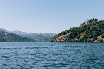 Naklejka premium Wide view of Lake Skadar with rippling blue water, forested hills and distant mountains under a clear sky, Skadar Lake National Park, Montenegro