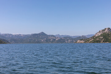 Fototapeta premium Wide panoramic view across Lake Skadar with gentle ripples, rocky green mountains and clear blue sky on a sunny day, Montenegro natural landscape travel background
