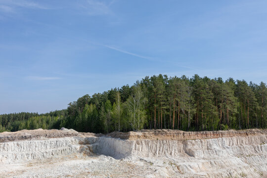 Wide landscape view of the Ural Bali kaolin quarry with bright white clay terraces, sandy ground and a dense pine forest line under a clear blue sky in Russia