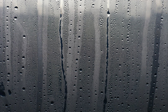 Numerous condensation droplets and wet streaks create an abstract pattern on a transparent glass surface. The moisture texture conveys a rainy weather atmosphere, cold, or high indoor humidity, formin
