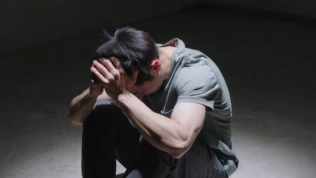 Distressed young man sitting on floor in dark room. Mental health concept with person holding head in hands. Static wide shot.