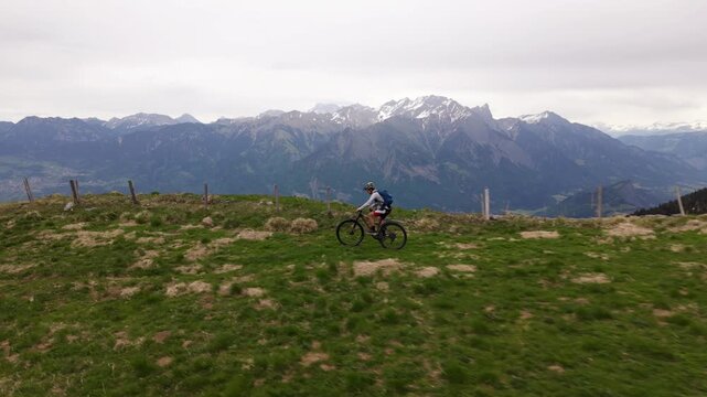 Drone side tracking footage of a mountain biker riding along a dramatic alpine ridge with steep drop-offs, remaining snow patches and panoramic mountain scenery during spring outdoor adventure.