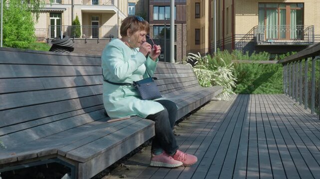 Elderly female sits on a wooden bench, changing sunglasses for eyeglasses for vision correction to see a smartphone screen. Struggling with presbyopia or age-related farsightedness. Senior health.
