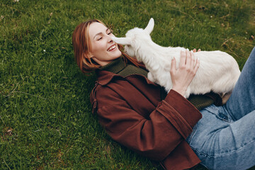 Fototapeta premium Relaxed young woman lying on grass, softly cuddling a white goat kid, enjoying a gentle outdoor moment, casual jacket and jeans, warm sunlight, peaceful countryside lifestyle scene.
