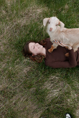 Fototapeta premium Casual lifestyle scene shows a woman lying on grass outdoors, a young goat kid perched on her chest, sharing an affectionate moment amid meadow, showcasing simple joy and bond with animals in a