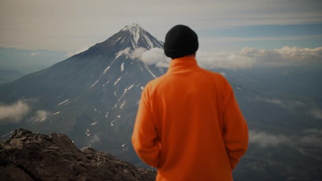 Young man in an orange jacket and black knitted hat stands on mountaintop and admiring volcano with its snow-capped peak in clouds. Back view. Slow motion. Concept of silence and pacification