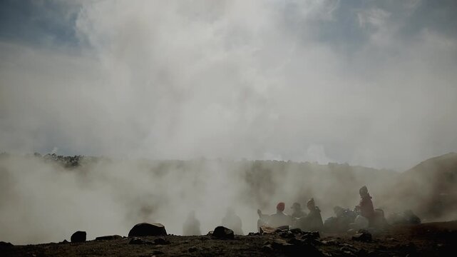 Kamchatka, Russia - August 24, 2025: Group of young people sit on edge of volcano's crater in smoke rising from cracks in solidified lava. Back view. Slow motion. Concept power of wild nature