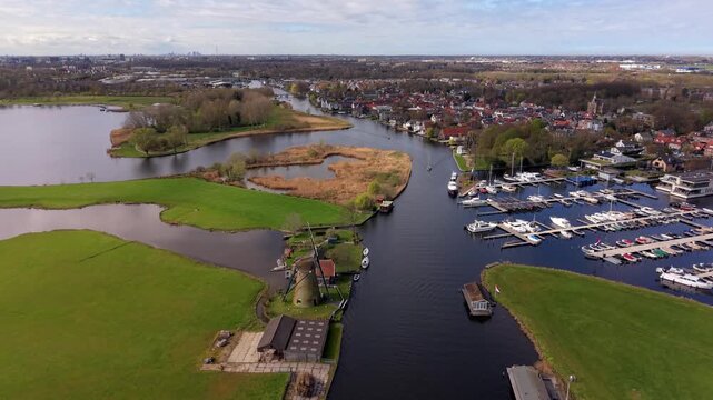 Aerial view of a traditional Dutch windmill by a river canal near a marina and village. Scenic landscape of rural Netherlands with green fields, waterways, and boats under a bright sky.