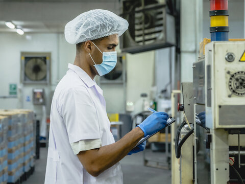 male quality control specialist in protective clothing operating machinery and holding a clipboard to ensure precise manufacturing standards while adhering to strict safety protocols.