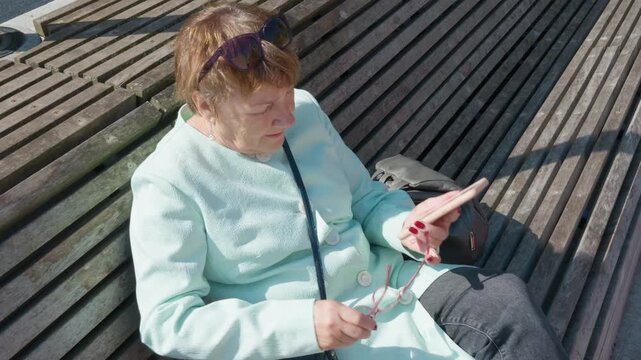 Senior woman with poor vision putting on reading glasses to see mobile screen. Elderly lady struggling with presbyopia or farsightedness while using smartphone. Vision correction and aging.