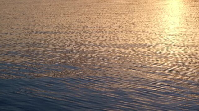 Close-up of water surface at sunset or sunrise, waves reflect golden sunlight, creating an abstract pattern of motion and light
